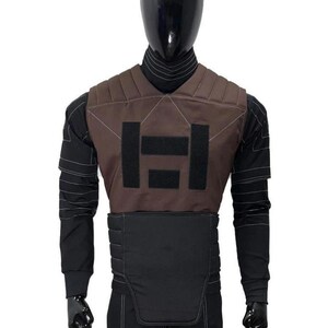 May include: A brown and black tactical vest worn over a black long-sleeved shirt with white stitching details. The vest features a brown upper chest panel with two black rectangular patches and a black padded lower section with horizontal stitching.