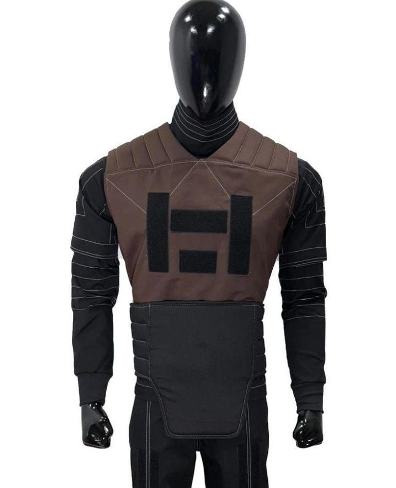 May include: A brown and black tactical vest worn over a black long-sleeved shirt with white stitching details. The vest features a brown upper chest panel with two black rectangular patches and a black padded lower section with horizontal stitching.