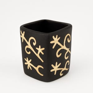 May include: A square, black ceramic container with a matte finish. The container is adorned with a repeating pattern of light beige, stylized floral and geometric designs. The interior is also black.