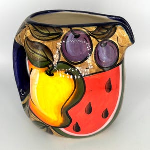 May include: A ceramic pitcher featuring a hand-painted design of a bright yellow pear, a slice of red watermelon with black seeds, and two purple plums. The pitcher has a dark blue handle and a dark blue rim, with green leaves and a tan background.