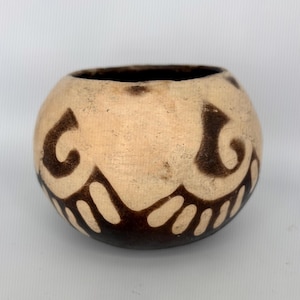 May include: A small, round, beige ceramic bowl with dark brown, stylized, curved patterns around the exterior. The bowl has a dark brown rim and base. The design is symmetrical, with the patterns resembling stylized swirls and teardrops.
