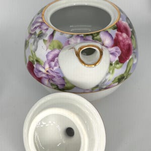 May include: A white porcelain teapot with a floral design featuring purple and red flowers on a light purple background. The teapot has a gold trim and a matching lid. The handle and spout are also white with gold accents. The lid is off to the side.