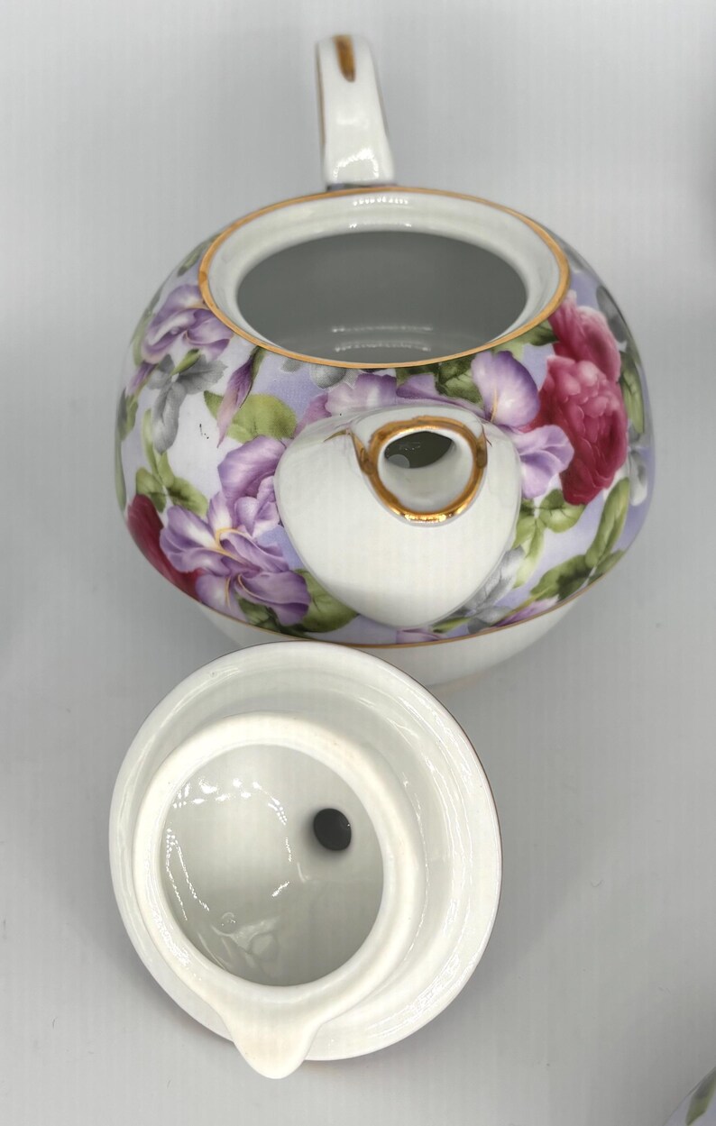May include: A white porcelain teapot with a floral design featuring purple and red flowers on a light purple background. The teapot has a gold trim and a matching lid. The handle and spout are also white with gold accents. The lid is off to the side.
