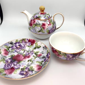 May include: A floral tea set featuring a teapot, teacup, and saucer. The set has a lavender background with purple and pink flowers and gold trim. The teapot has a gold knob on the lid and a curved spout. The teacup has a handle.