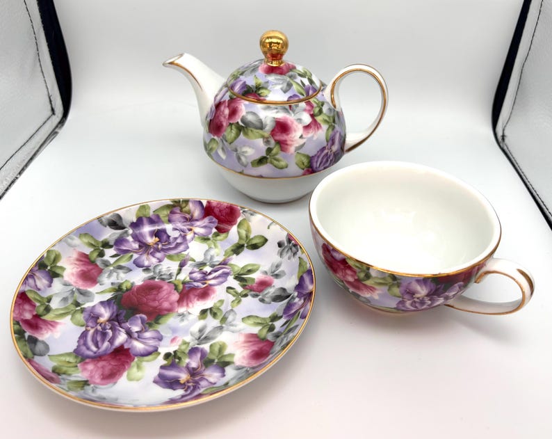 May include: A floral tea set featuring a teapot, teacup, and saucer. The set has a lavender background with purple and pink flowers and gold trim. The teapot has a gold knob on the lid and a curved spout. The teacup has a handle.