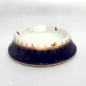 May include: A round porcelain ashtray featuring a deep navy blue base with a gold scalloped trim and a white interior. The rim is decorated with a delicate gold pattern, and the bowl includes two notches for resting items on the edge.