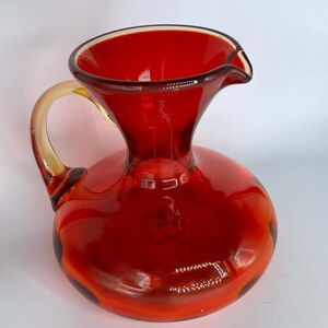 May include: A vibrant red glass pitcher with a rounded body and a curved handle. The pitcher has a spout for easy pouring and a yellow-tinted handle. This decorative pitcher is ideal for serving beverages.