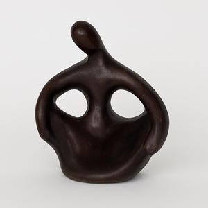 May include: A dark brown, abstract sculpture of a human figure. The sculpture has a rounded body with two large, oval openings and a slender head. The figure appears to be in a seated or reclining position, with arms extending outward.