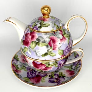 May include: A floral tea set featuring a teapot, teacup, and saucer. The set is decorated with pink, purple, and white roses and green leaves on a light purple background. The rims are trimmed with gold. The teapot has a gold knob on the lid.