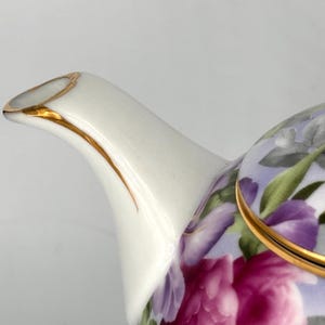 May include: Close-up of a white porcelain teapot with a gold-trimmed spout and lid. The teapot is decorated with a floral pattern featuring pink and purple flowers and green leaves. The design is set against a light purple background.