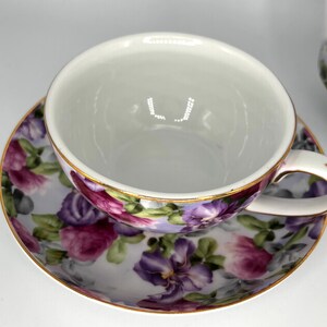 May include: White teacup and saucer set with a floral design. The cup and saucer feature a pattern of purple, pink, and green flowers on a light background. The rims are trimmed with gold.