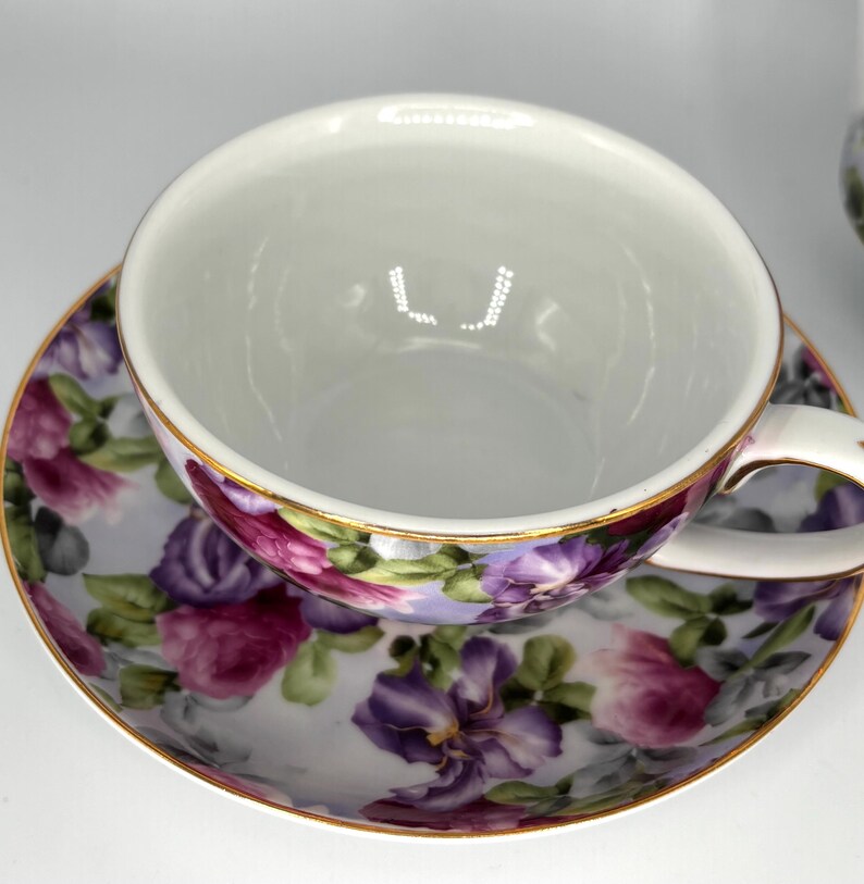 May include: White teacup and saucer set with a floral design. The cup and saucer feature a pattern of purple, pink, and green flowers on a light background. The rims are trimmed with gold.