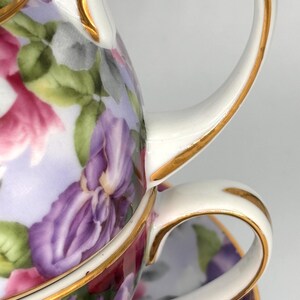 May include: A white porcelain teacup and teapot set with a floral design featuring purple irises, pink flowers, and green leaves. The rims and handles are trimmed with gold. The set is designed for tea parties.