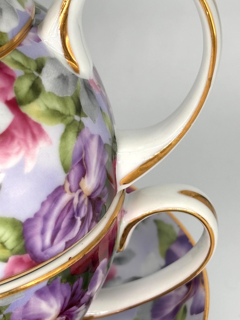 May include: A white porcelain teacup and teapot set with a floral design featuring purple irises, pink flowers, and green leaves. The rims and handles are trimmed with gold. The set is designed for tea parties.