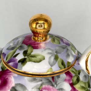 May include: Close-up of a floral teapot with a gold-colored lid and handle. The teapot features a design of pink and purple roses with green leaves on a light purple background. The rim and handle are trimmed with gold.