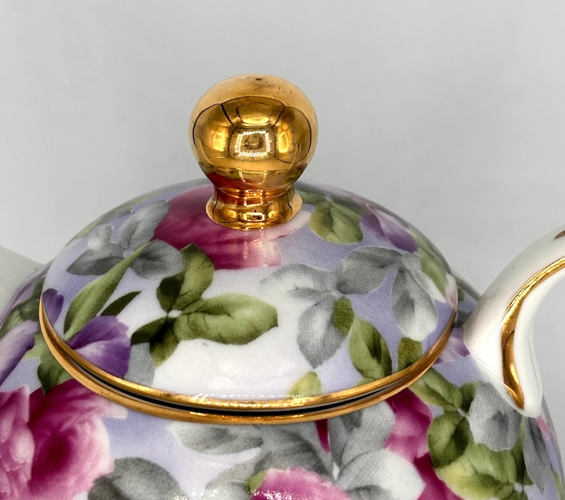 May include: Close-up of a floral teapot with a gold-colored lid and handle. The teapot features a design of pink and purple roses with green leaves on a light purple background. The rim and handle are trimmed with gold.