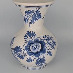 May include: A ceramic vase with a bulbous body and a flared rim, featuring a white base color decorated with hand-painted blue floral patterns and leafy sprigs. The design includes a large central flower surrounded by foliage and thin horizontal lines.