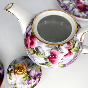 May include: A white porcelain teapot with a floral design featuring pink and purple roses, green leaves, and gold trim. The lid is off, revealing the interior. A matching saucer is visible in the background. A classic tea set.