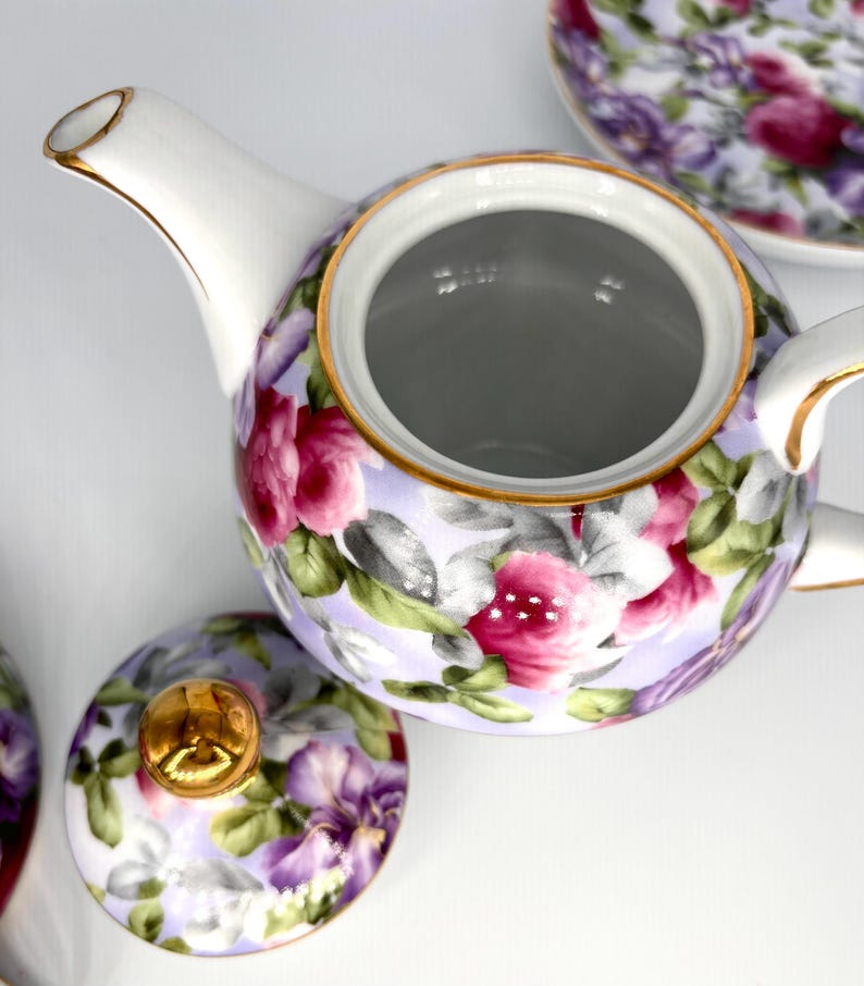 May include: A white porcelain teapot with a floral design featuring pink and purple roses, green leaves, and gold trim. The lid is off, revealing the interior. A matching saucer is visible in the background. A classic tea set.