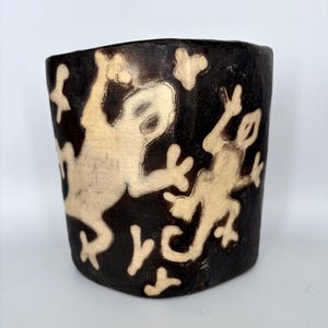 May include: A dark brown, cylindrical ceramic vase with a cream-colored design of two stylized frogs. The frogs are depicted in a playful pose, with their limbs outstretched. The vase has a slightly textured surface.