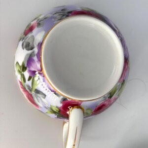 May include: A white teacup with a floral design. The cup's exterior features a lavender background with pink, red, and gray flowers and green leaves. The rim and handle are trimmed with gold.
