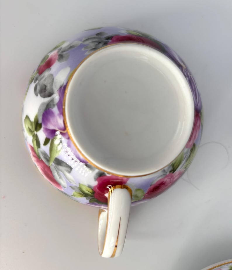 May include: A white teacup with a floral design. The cup's exterior features a lavender background with pink, red, and gray flowers and green leaves. The rim and handle are trimmed with gold.