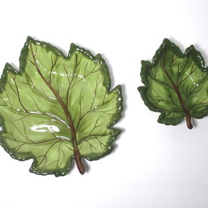 May include: Two ceramic dishes shaped like grape leaves, one larger than the other. Both feature a glossy finish with light green centers, darker green edges, and prominent brown veins radiating from the stem base to the serrated leaf margins.