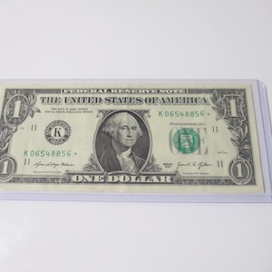 May include: A United States one dollar Federal Reserve note featuring a portrait of George Washington, enclosed in a clear plastic protective sleeve. The bill displays the serial number K06548856 with a star symbol and green treasury seal.