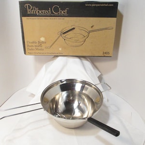 May include: A stainless steel double boiler set from The Pampered Chef. The set includes a large, shiny bowl with a handle and a long, thin handle. The box in the background has the product name and a diagram.