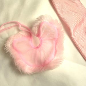Könnte beinhalten: Ein rosa herzförmiges Accessoire mit flauschigem, federartigem Besatz und einem durchsichtigen, tüllartigen Mittelteil. Ein passendes rosa Band ist ebenfalls sichtbar. Die Artikel sind vor einem weißen Hintergrund platziert.