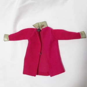 May include: A bright pink doll-sized coat with a gold-colored collar and cuffs. The coat is open in the front and has a single button closure. The sleeves are extended, showcasing the design.