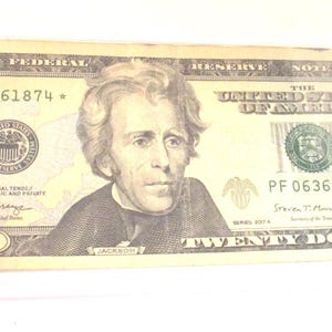 Pode incluir: Uma nota de vinte dólares em um suporte de plástico transparente. A nota apresenta um retrato de Andrew Jackson e o texto "Twenty Dollars". O número de série é visível, juntamente com o texto "The United States of America".