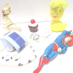 May include: A collection of items including a white goose figurine, a chocolate cupcake with a red cherry, a yellow duck container with hair, a blue and white Avon box, a beaded necklace with blue accents, and a Superman figurine in a blue suit.
