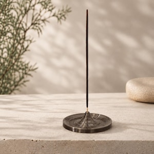 May include: A dark grey, circular incense holder with a conical centre piece that holds a single, thin, dark brown incense stick upright. The holder features a smooth, polished finish with subtle, swirling patterns throughout the material.