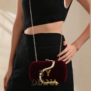 May include: A burgundy velvet clutch with a gold snake design and chain strap. The clutch has the text "BY DIANA" in gold. The bag is held by a person wearing a black dress.