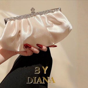 Könnte beinhalten: Cremefarbene Satin-Clutch mit silbernem Verschluss und Strassbesatz. Die Clutch wird in einer Hand gehalten, mit dem Text "BY DIANA" in goldfarbenen Buchstaben. Die Clutch hat ein gerafftes Design und einen dekorativen Verschluss.
