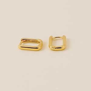 May include: Two gold-colored, rectangular hoop earrings. The earrings have a smooth, polished finish and a simple, modern design. The earrings are displayed on a neutral background.