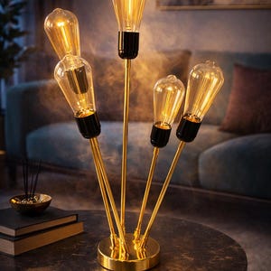 May include: A modern gold-toned table lamp with five exposed light bulbs. The bulbs are clear with a warm, amber glow. The lamp has a circular base and slender, upward-reaching stems. The lamp is on a dark, round table.