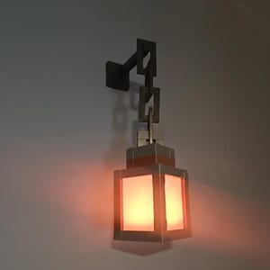 May include: A wall-mounted lantern with a rectangular, illuminated shade. The lantern is suspended by a chain-like structure and a square mounting plate. The light emits a warm, orange glow, creating a cozy ambiance. The fixture is made of metal.
