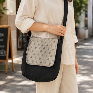 May include: A black crossbody bag featuring a rectangular flap with a light beige fabric overlay. The flap is decorated with a repeating pattern of small, dark, four-pointed geometric shapes. The bag includes a long, adjustable black shoulder strap.