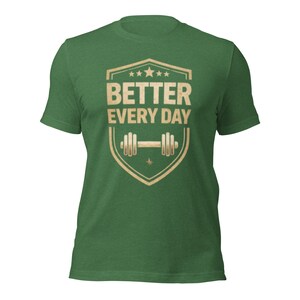 May include: Green t-shirt with a shield-shaped graphic in tan and white. The graphic features the text "BETTER EVERY DAY" above a barbell illustration. The shirt is made of a soft, textured fabric.