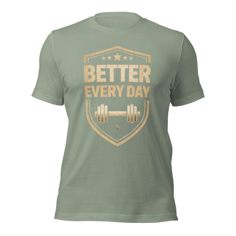 May include: Sage green t-shirt with a distressed gold graphic. The graphic features the text "BETTER EVERY DAY" inside a shield shape, with a dumbbell icon below. The shirt is a short-sleeved crew neck.