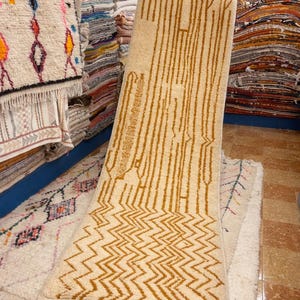 May include: A beige rectangular rug with a geometric pattern. The rug features a zig-zag design at the bottom and vertical lines in the center, all in a warm brown color. The rug is displayed in a shop with other rugs.