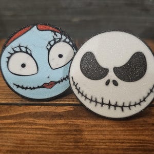 May include: Two circular decorations featuring characters from The Nightmare Before Christmas. One is light blue with a stitched smile and red lips, the other is white with a stitched smile and black glitter eyes.