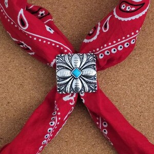 May include: A red bandana with a white paisley pattern, secured with a silver-colored bolo tie featuring a turquoise stone center. The bandana is folded and tied, creating a stylish accessory.