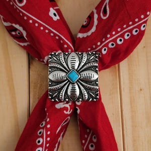 May include: A red bandana with a white paisley pattern is held together by a silver-toned square bolo tie clasp. The clasp features a turquoise stone at the center, surrounded by a floral design.