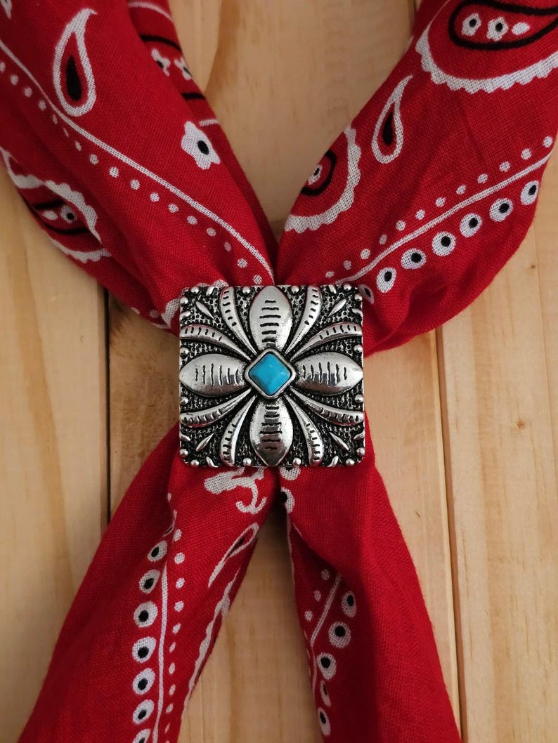 May include: A red bandana with a white paisley pattern is held together by a silver-toned square bolo tie clasp. The clasp features a turquoise stone at the center, surrounded by a floral design.
