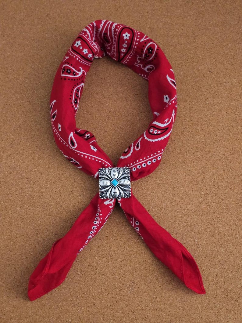 May include: A red bandana with a white paisley pattern, secured with a silver-colored bolo tie featuring a turquoise stone center. The bandana is folded and tied, creating a stylish accessory.