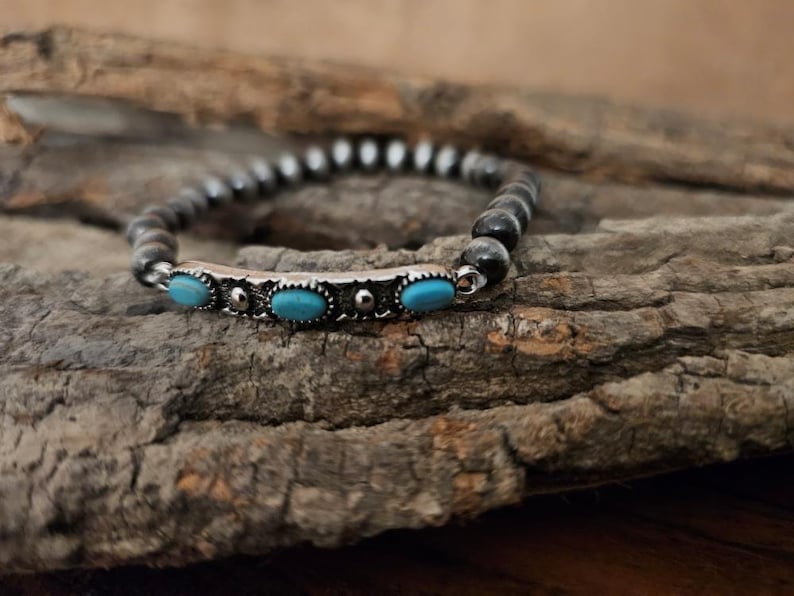 May include: A beaded bracelet with a silver accent featuring three oval turquoise stones. The bracelet is made of dark gray beads and is displayed on a piece of textured wood.