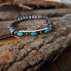 May include: A beaded bracelet with a silver bar featuring three oval turquoise stones. The bracelet is made of small, round, silver-colored beads. The jewelry is displayed on a piece of textured, brown wood.
